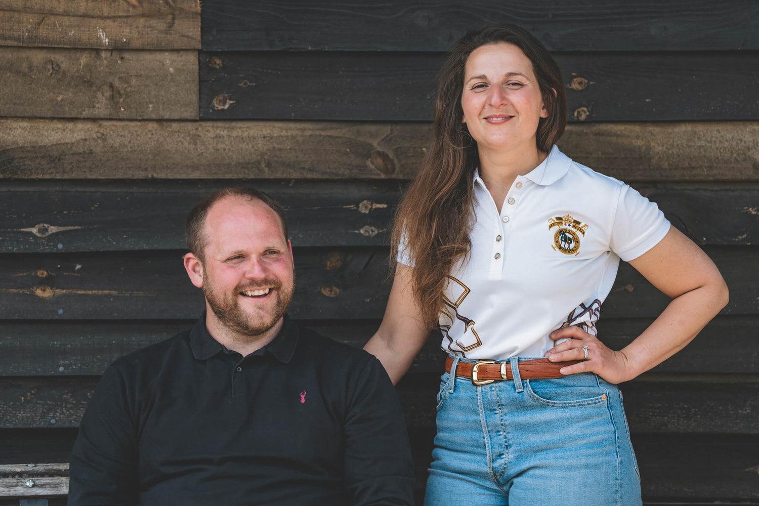 Melanie and Chris, owners of Total Contact Saddles. Chris is sitting on a bench while Melanie stands next to him
