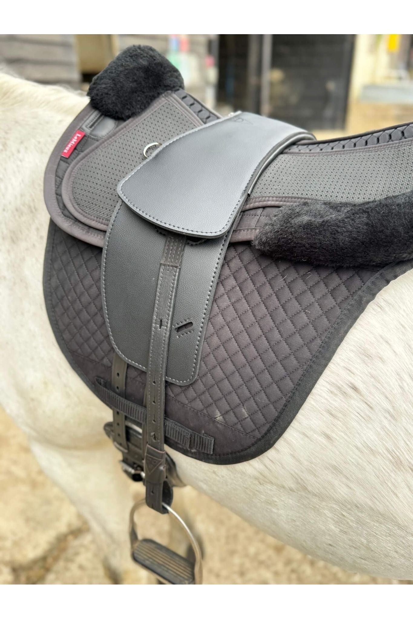 A synthetic black long billet Total Contact Saddle on a black saddle pad and a black half pad on a grey pony