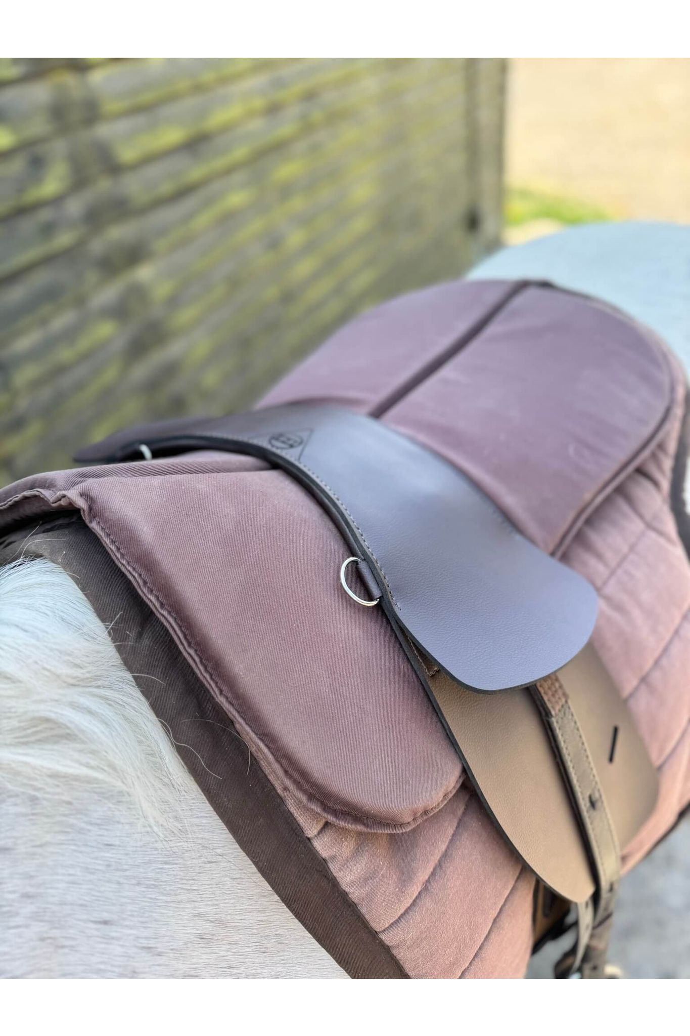 A synthetic chocolate brown treeless Total Contact Saddle on a brown saddle pad and a brown half pad on a grey pony