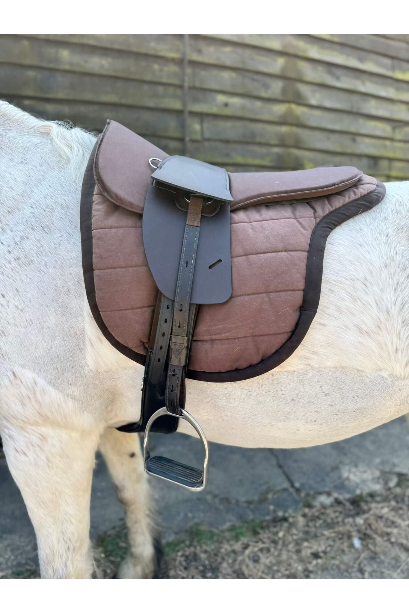A synthetic chocolate brown treeless Total Contact Saddle on a brown saddle pad and a brown half pad on a grey pony