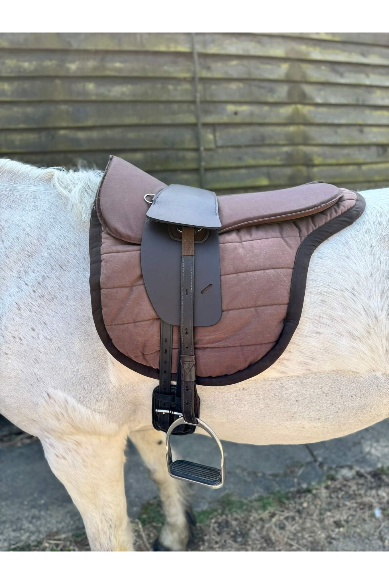 A synthetic chocolate brown treeless Total Contact Saddle on a brown saddle pad and a brown half pad on a grey pony