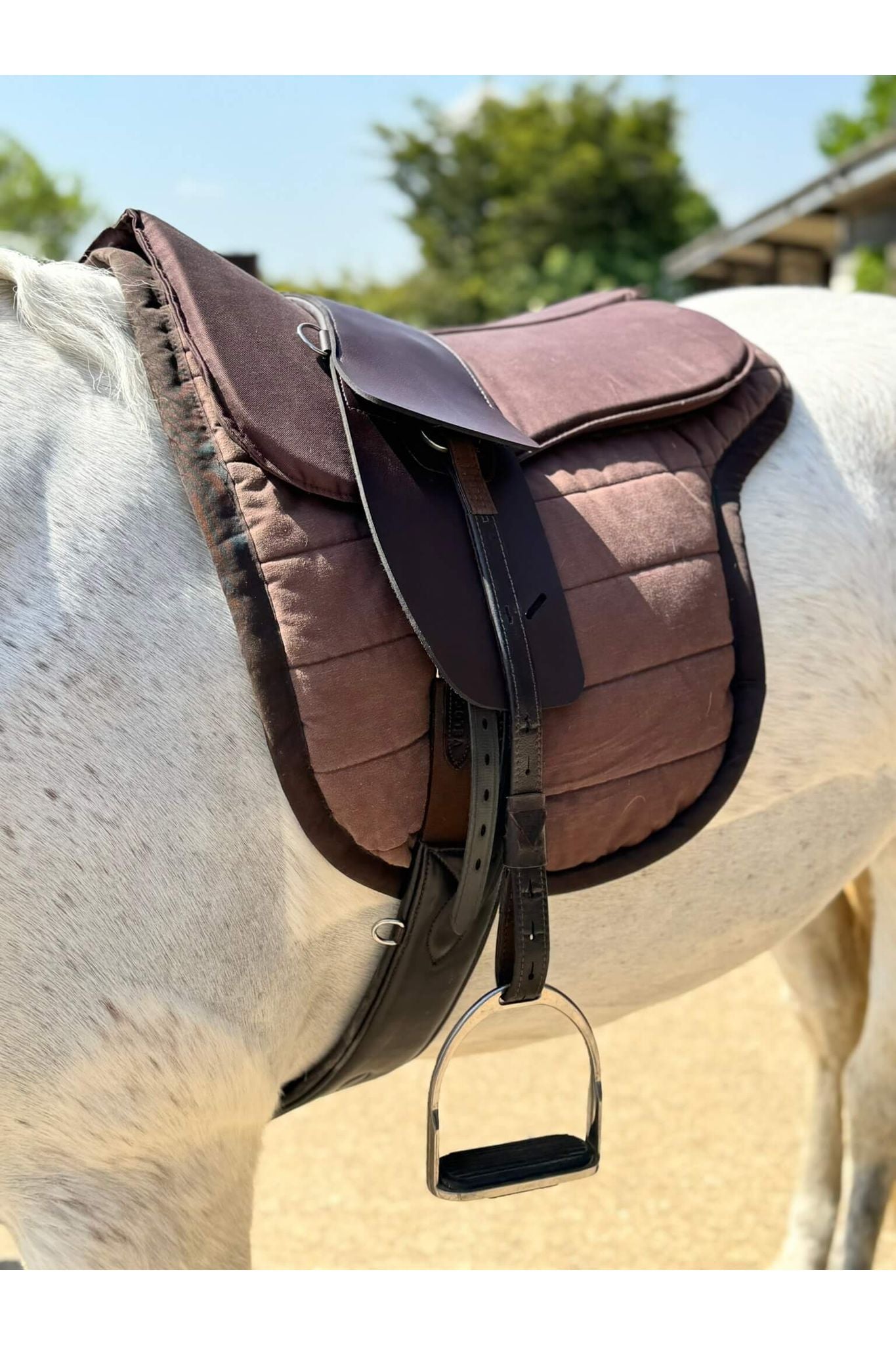 A synthetic chocolate brown treeless Total Contact Saddle on a brown saddle pad and a brown half pad on a grey pony standing in the sun