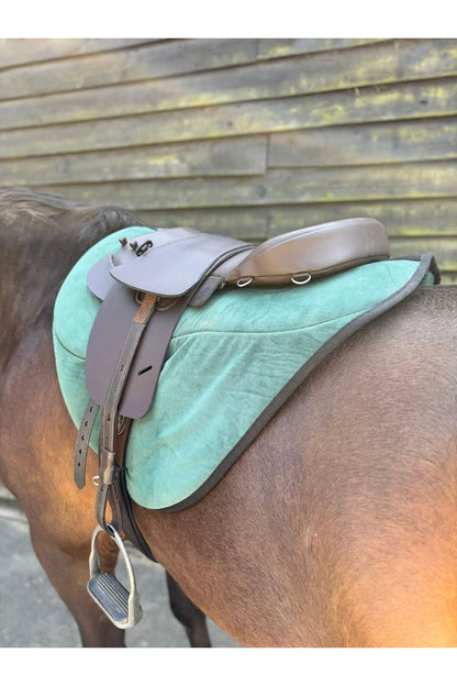 A brown synthetic treeless total contact saddle and a brown synthetic saddle seat pad on a dark bay horse wearing a green saddle pad